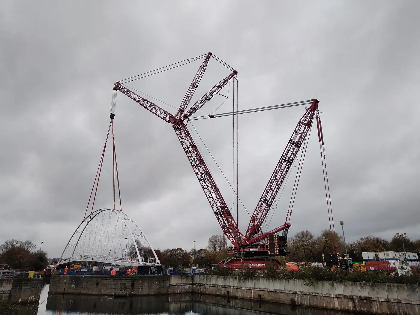 A red lattice-boom crawler crane lifting an arched footbridge span over a river