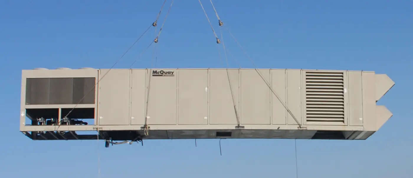 A rooftop air-handling unit hanging from a crane's rigging against a clear sky