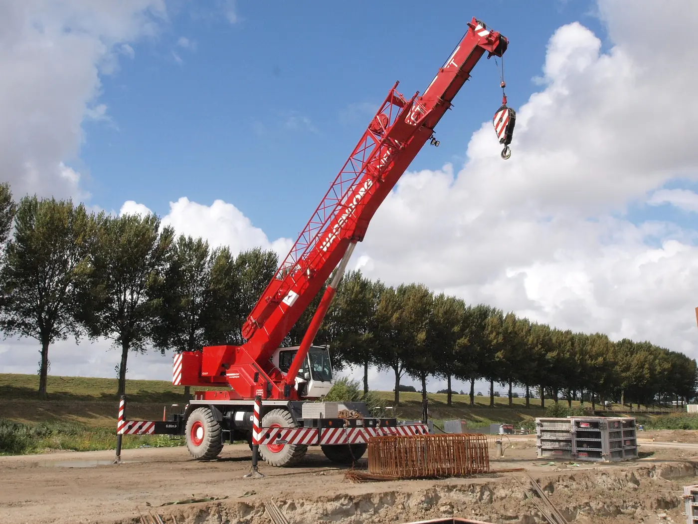 A red rough-terrain crane with its boom raised and hook block hanging, set up on a gravel pad